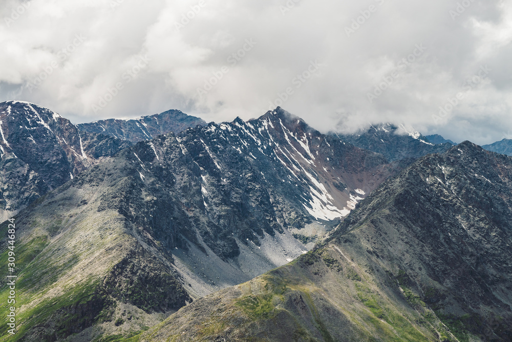 Atmospheric alpine landscape with big mountain ridge and snowbound peak in low clouds. Cloudy sky over huge mountain range. Giant snowy craggy rocks and rocky valley. Majestic scenery on high altitude Atmospheric alpine landscape with big mountain ridge and snowbound peak in low clouds. Cloudy sky over huge mountain range. Giant snowy craggy rocks and rocky valley. Majestic scenery on high altitude