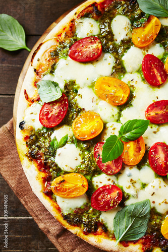 Pizza with mozzarella cheese, red and yellow cherry tomatoes, olive pesto and fresh basil. Home made food. Concept for a tasty and hearty meal. Rustic wooden background. Top view. 