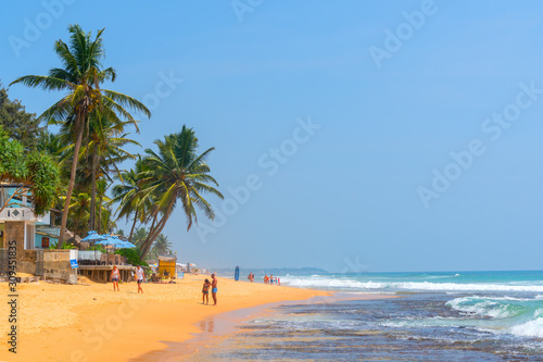 Hikkaduwa, Sri Lanka. March 6, 2018. Sunny Beach on Indian Ocean. Palm trees on sandy shore. People on coast.