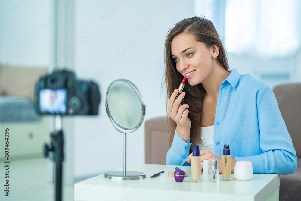 Happy young smiling woman blogger using red lipstick for paints lips during recording her beauty blog about makeup and cosmetics at home. Blogging and freelance people Happy young smiling woman blogger using red lipstick for paints lips during recording her beauty blog about makeup and cosmetics at home. Blogging and freelance people