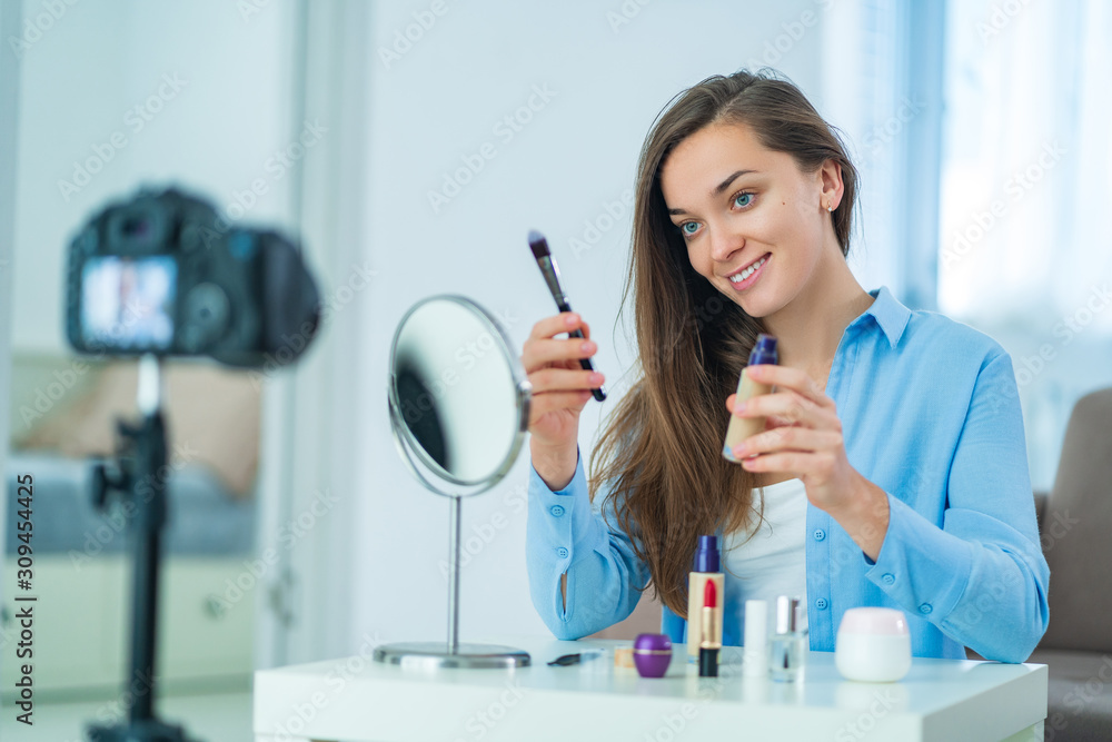 Happy young smiling woman video blogger influencer holding brush and foundation during recording her beauty blog about makeup and cosmetics at home. Blogging and influencing on the audience Happy young smiling woman video blogger influencer holding brush and foundation during recording her beauty blog about makeup and cosmetics at home. Blogging and influencing on the audience