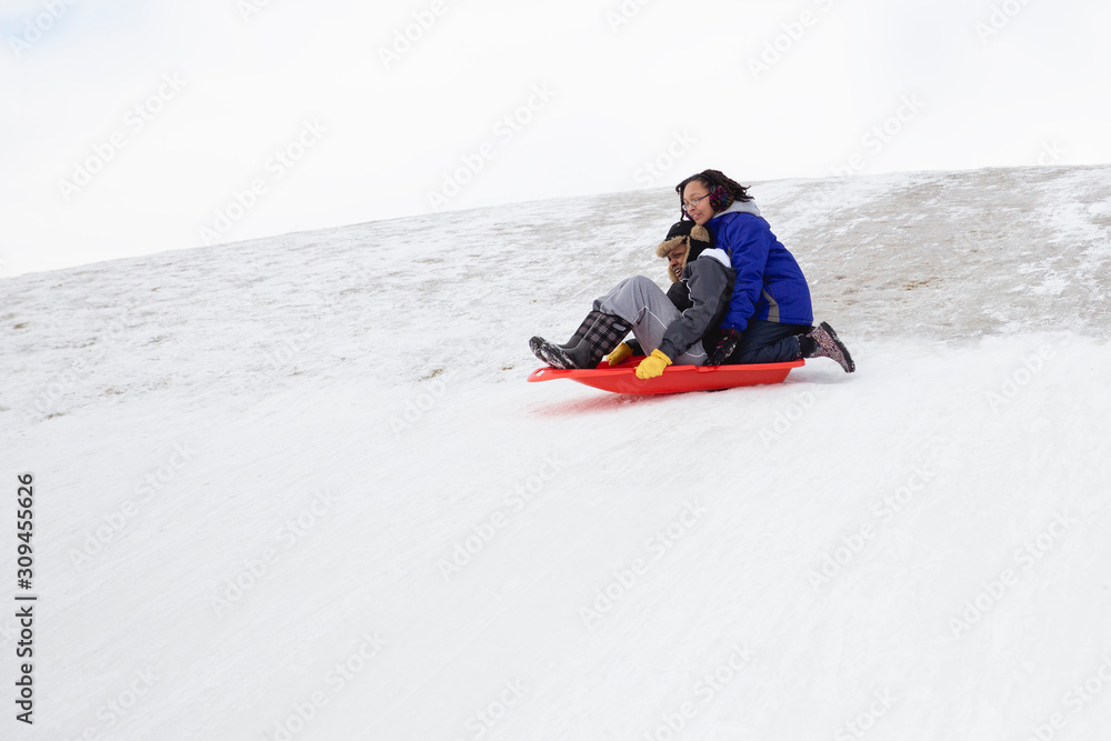 Black mother with her mixed race teen happily tobogganing down a hill together Black mother with her mixed race teen happily tobogganing down a hill together