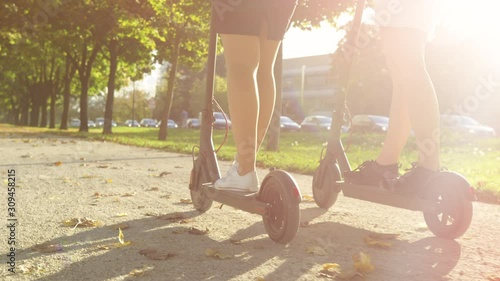 SLOW MOTION, LOW ANGLE, CLOSE UP, DOF: Golden sunbeams shine on two unrecognizable women ride high tech scooters around the park on a sunny autumn day. Friends enjoy a scenic scooter ride around park