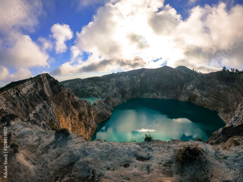 View on the Kelimutu volcanic crater lakes in Flores, Indonesia. Lakes are shining with many shades of turquoise and blue. Sun shines through clouds. Barren and sharp slopes of the volcanic crater © Chris