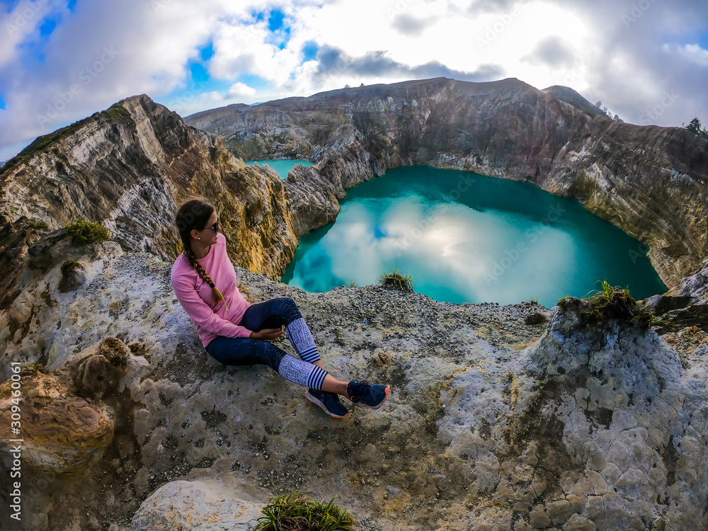 Woman sitting at the volcano rim and watching the Kelimutu volcanic crater lakes in Moni, Flores, Indonesia. Woman is relaxed and calm, enjoying the view on lake shining with many shades of turquoise