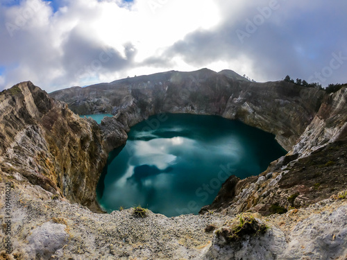 View on the Kelimutu volcanic crater lakes in Flores, Indonesia. Lakes are shining with many shades of turquoise and blue. Sun shines through clouds. Barren and sharp slopes of the volcanic crater