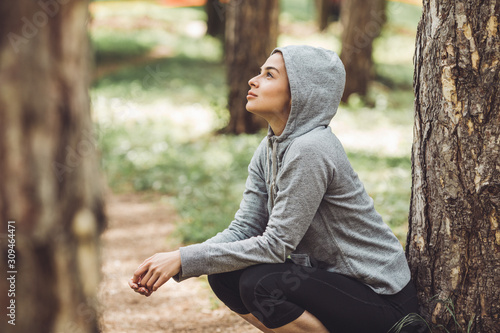 A young woman resting from jogging in the park by a tree. She is wearing a grey hoodie and gazes upwards, reflecting.