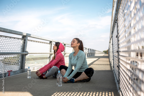 Two young women in sportswear preparing for jogging. They are sitting and stretching legs in yoga cobbler's pose on pedestrian bridge.