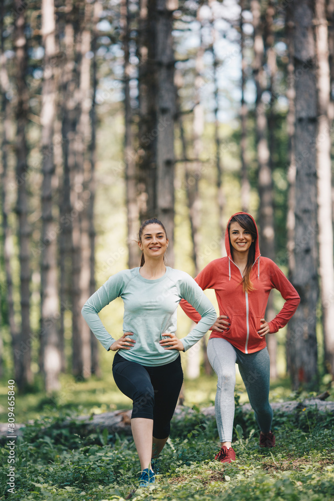 Fototapeta premium Two beautiful young women in sportswear doing lunges in woods.