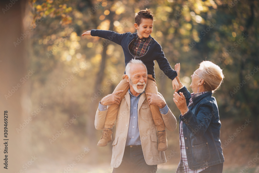 Grandparents having a lovely autumn day with their grandson in nature. The grandfather is carying his grandson on his shoulders while the grandmother holds the child's hands. - obrazy, fototapety, plakaty 