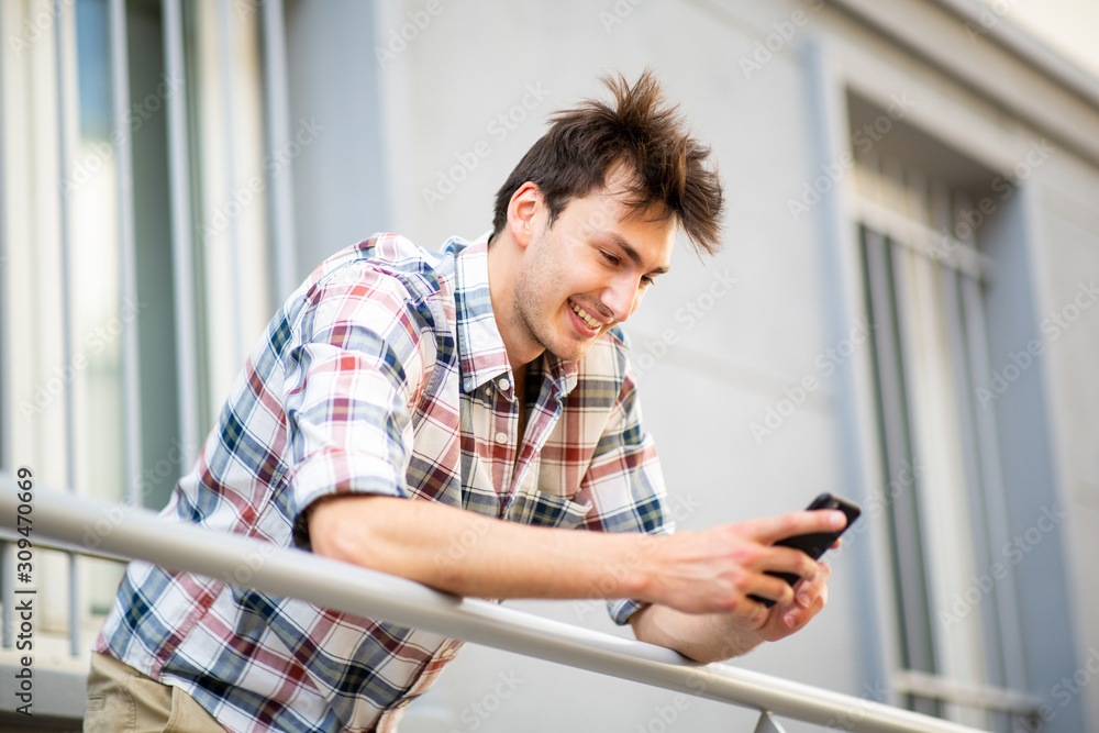 smiling young man on balcony looking at cellphone - obrazy, fototapety, plakaty 