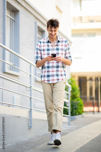Full body happy young man walking and looking at mobile phone