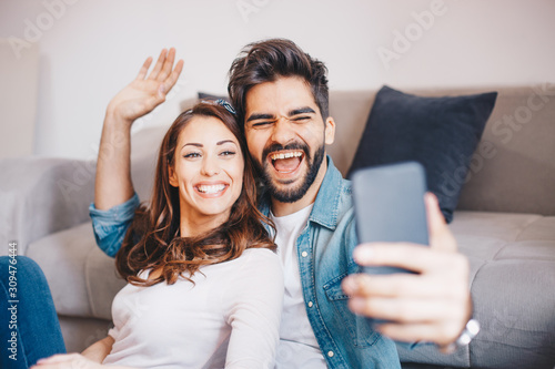 Beautiful young Caucasian couple smiling and taking a selfie while they are sitting by the couch and waiting for a tv show to start in their living room.