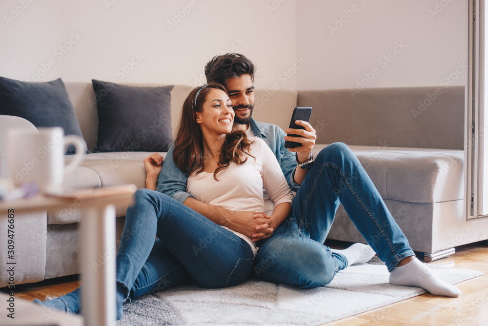 Cute Caucasian newly weds watching photos on smart-phone while they are embracing and relaxing on the floor by the couch.