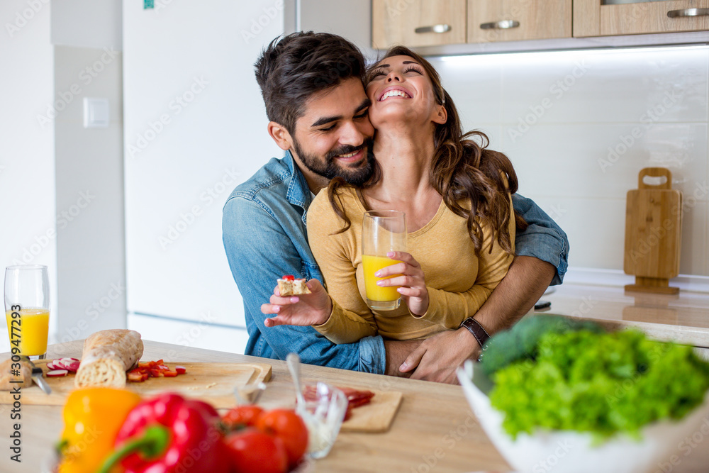 Young handsome Caucasian man hugging his beautiful cheerful girlfriend with love while they are having breakfast in the kitchen.