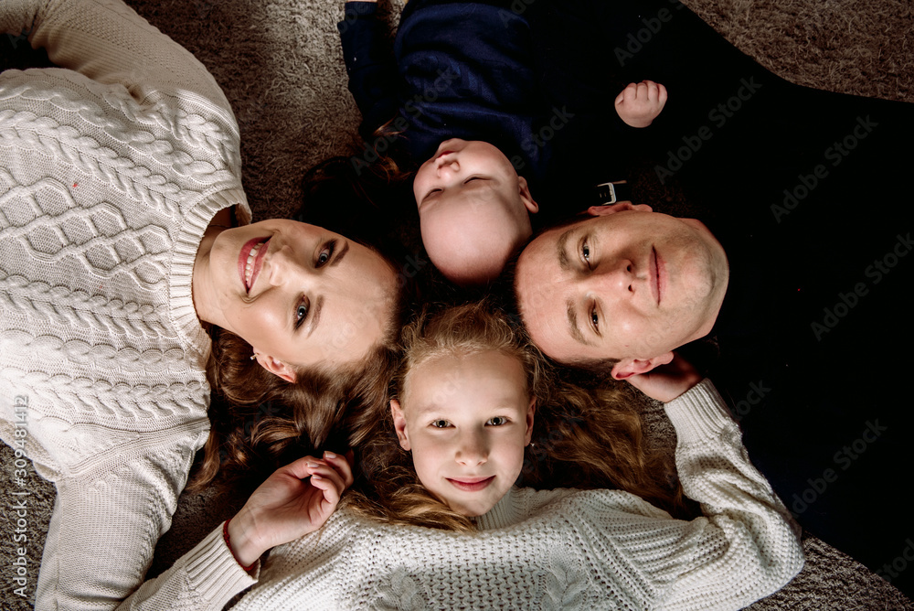 Top view of beautiful young parents, their cute little daughter and son holding hands, looking at camera and smiling, lying on wooden floor