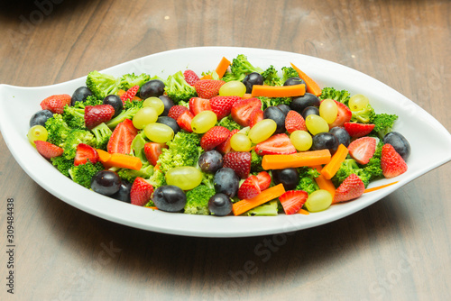 Closeup of fruit salad with broccoli, green and black grapes, strawberry slices and sliced ​​carrots and cubes. Delicious, nutritious and healthy meal in white ceramic serving dish on the wooden table