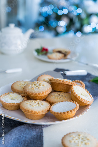 Traditional english festive pastry mince pies on served for tea time table with lightened christmas tree on background. Cozy home mood. Vertical card. Close up, selective focus. Copy space.