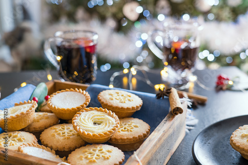 Warm cosy composition of traditional english festive pastry mince pies in wooden tray with blurred background of mulled wine drinks, lights garland and christmas tree on background. Close up.