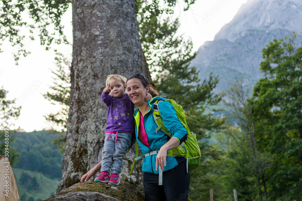 Mother supporting her daughter standing on tree trunk exploring the forest view in mountains.