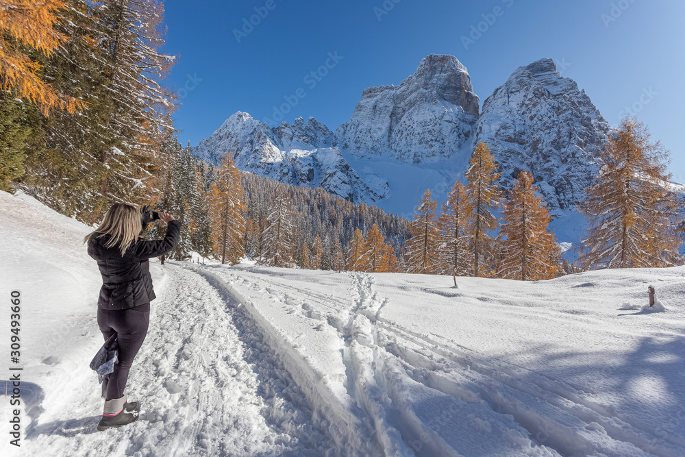 Blonde girl takes a picture with the phone at a beautiful snow-covered Dolomite mountain surrounded by larch forests with autumn color. Concept: fun in the snow, winter landscape, snow holidays 