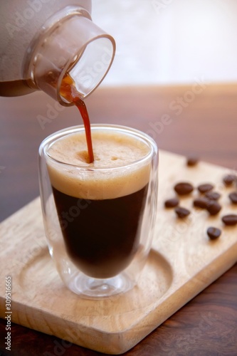 Shaken ice espresso shot pouring from shaker bottle in espresso glass shot served on wood plate with coffee beans background.