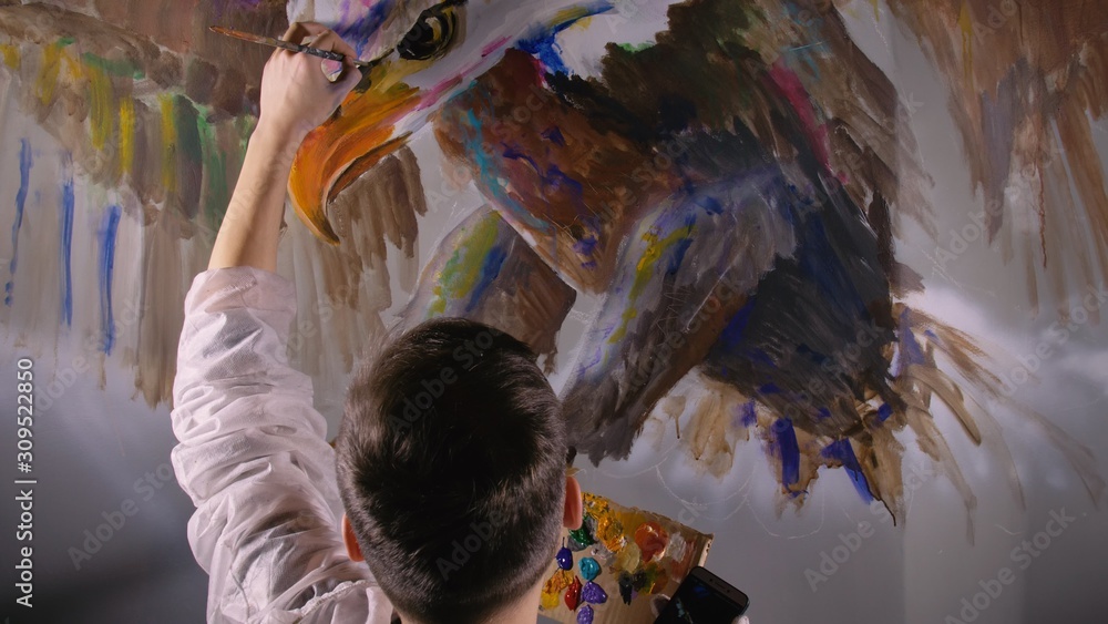Fototapeta premium Artist designer draws an eagle on wall. Craftsman decorator paints picture with acrylic oil color looking at sketch in phone. Painter painter dressed in paint coat. Indoor. Dark magic cinematic look.