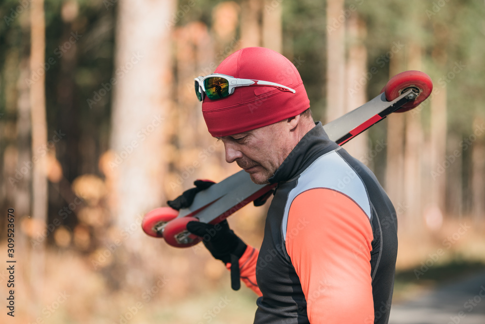 Fototapeta premium Training an athlete on the roller skaters. Biathlon ride on the roller skis with ski poles, in the helmet. Autumn workout. Roller sport. Adult man riding on skates.