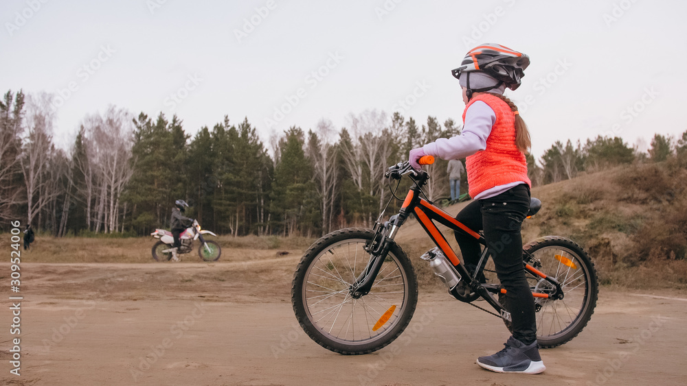 One caucasian children rides bike road track in dirt park. Girl riding black orange cycle in racetrack. Kid goes do bicycle sports. Biker motion ride with backpack and helmet. Mountain bike hardtail. One caucasian children rides bike road track in dirt park. Girl riding black orange cycle in racetrack. Kid goes do bicycle sports. Biker motion ride with backpack and helmet. Mountain bike hardtail.