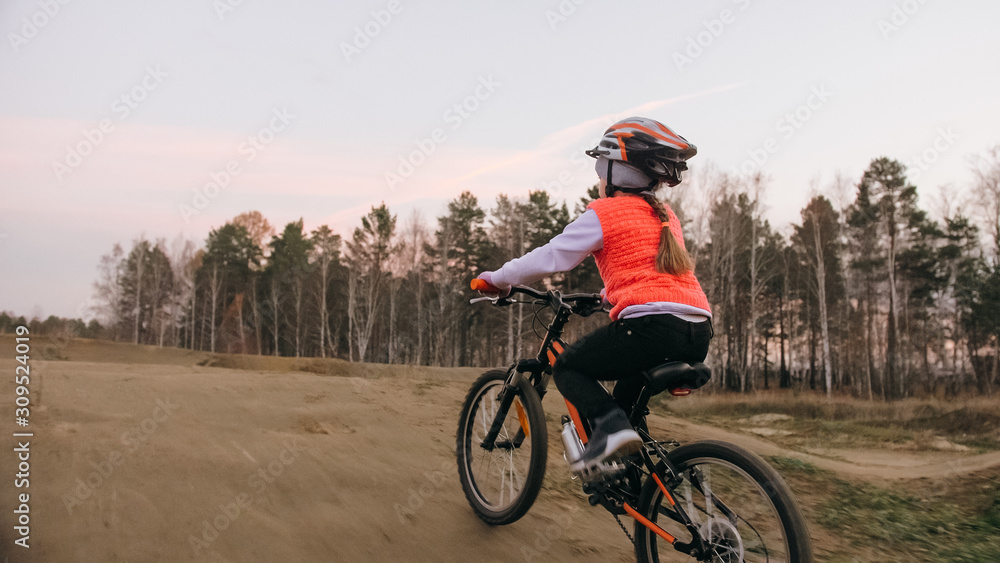 One caucasian children rides bike road track in dirt park. Girl riding black orange cycle in racetrack. Kid goes do bicycle sports. Biker motion ride with backpack and helmet. Mountain bike hardtail. One caucasian children rides bike road track in dirt park. Girl riding black orange cycle in racetrack. Kid goes do bicycle sports. Biker motion ride with backpack and helmet. Mountain bike hardtail.