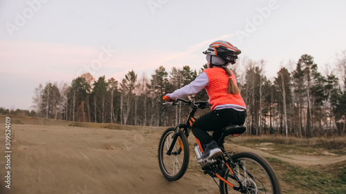 One caucasian children rides bike road track in dirt park. Girl riding black orange cycle in racetrack. Kid goes do bicycle sports. Biker motion ride with backpack and helmet. Mountain bike hardtail. © ivandanru