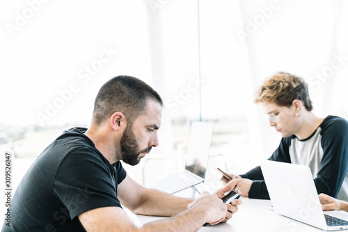 Two young men looking the mobile one to each side of the table in a workspace
