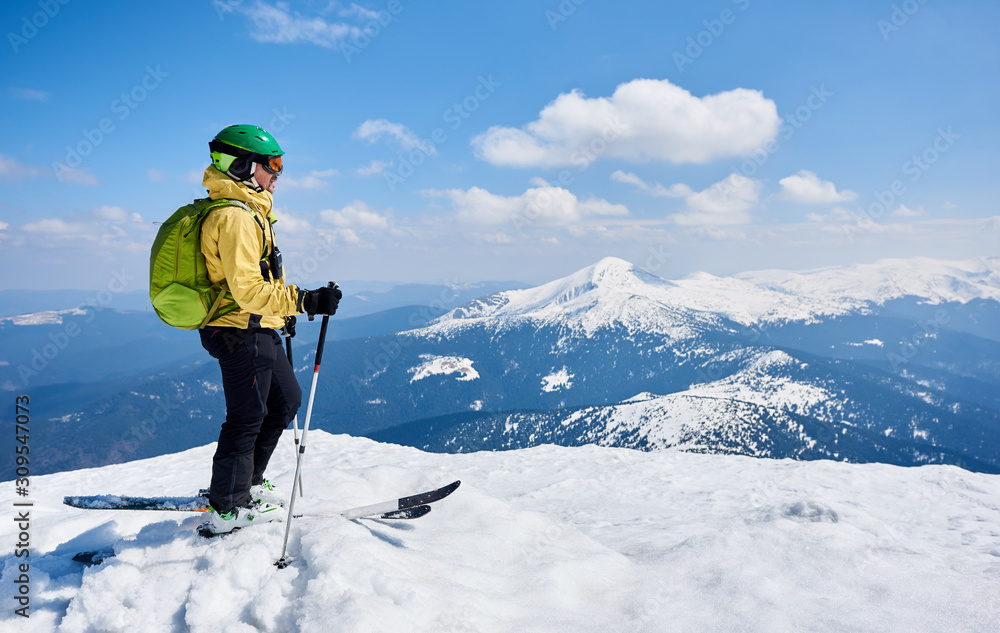 Sportsman skier in helmet and goggles with backpack standing in profile on skis holding ski poles in deep white snow, on copy space background of bright blue sky enjoying beautiful mountain view. Sportsman skier in helmet and goggles with backpack standing in profile on skis holding ski poles in deep white snow, on copy space background of bright blue sky enjoying beautiful mountain view.