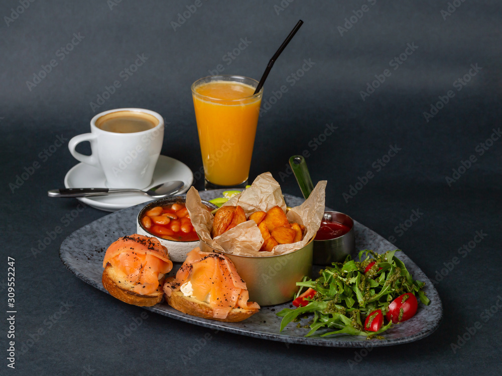 Hearty breakfast - fried potatoes, beans, two sandwiches with fetax cheese and salmon, rucola and tomato salad with pesto sauce. On a dark background Hearty breakfast - fried potatoes, beans, two sandwiches with fetax cheese and salmon, rucola and tomato salad with pesto sauce. On a dark background