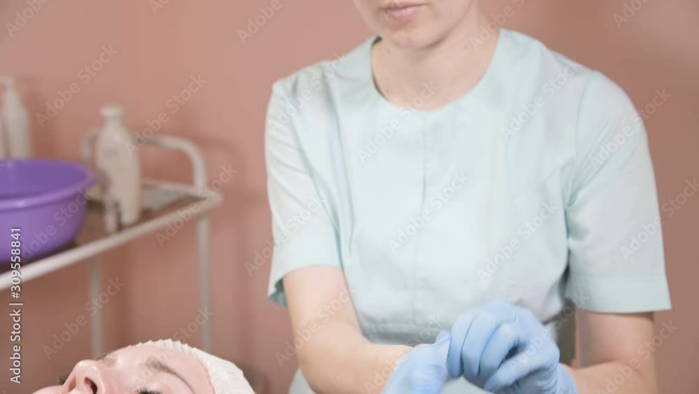 Professional woman beautician puts on her hands blue rubber clean sterile gloves before doing the procedure for cosmetic skin care of the client's face. The concept of professionalism in cosmetology Professional woman beautician puts on her hands blue rubber clean sterile gloves before doing the procedure for cosmetic skin care of the client's face. The concept of professionalism in cosmetology