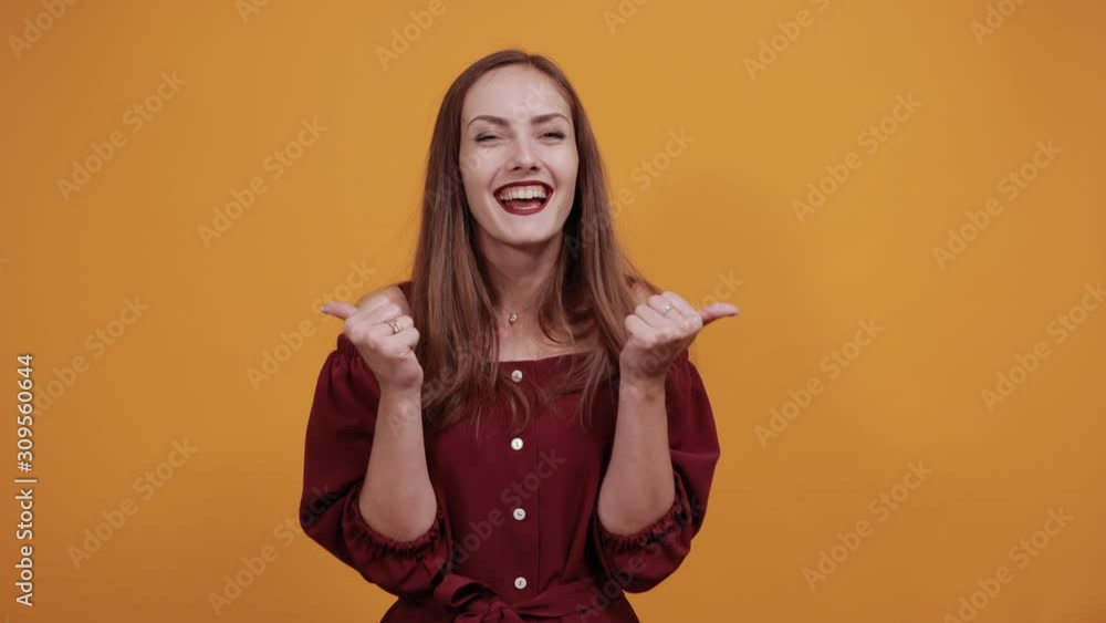 Charming caucasian young woman in maroon dress isolated on orange background in studio claps her hands, pointing thumbs aside. People sincere emotions, lifestyle concept. Charming caucasian young woman in maroon dress isolated on orange background in studio claps her hands, pointing thumbs aside. People sincere emotions, lifestyle concept.