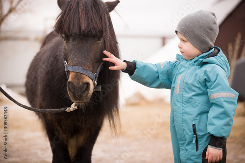 Cute Kid boy in turquoise overalls stroking an Icelandic pony with a funny forelock. Child thanks the horse after hippotherapy. Brown pony looking at the camera