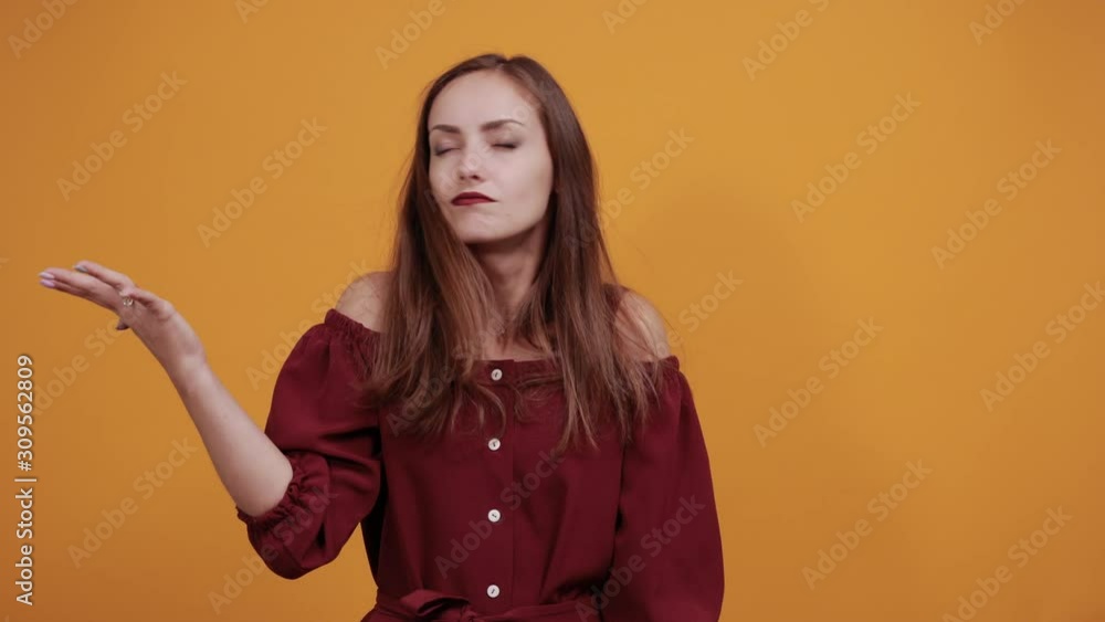 Brunette woman in maroon dress isolated on orange background in studio pointing palm aside, looking at camera. People sincere emotions, lifestyle concept. Brunette woman in maroon dress isolated on orange background in studio pointing palm aside, looking at camera. People sincere emotions, lifestyle concept.