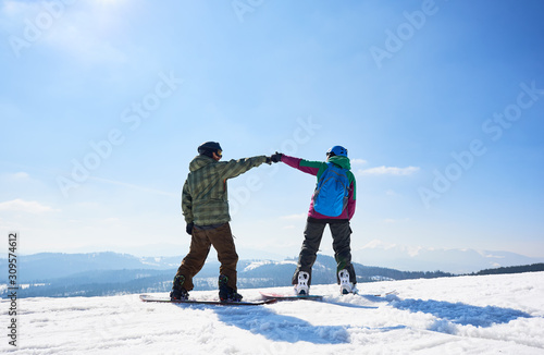 Back view of two happy tourists snowboarders on mountain summit on copy space background of blue sky and woody mountains on sunny winter day. Extreme winter sports, active lifestyle concept.