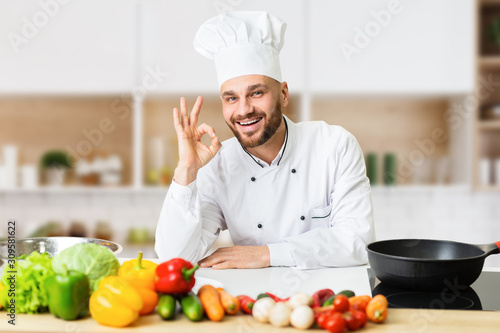 Smiling Cook Man Gesturing Okay Standing Near Table In Kitchen photo