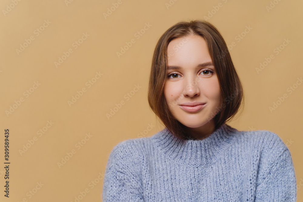 Headshot of attractive woman with long hair, charming look, wears purple knitted sweater, looks directly at camera, models over brown background, blank space on left side. People, tenderness Headshot of attractive woman with long hair, charming look, wears purple knitted sweater, looks directly at camera, models over brown background, blank space on left side. People, tenderness