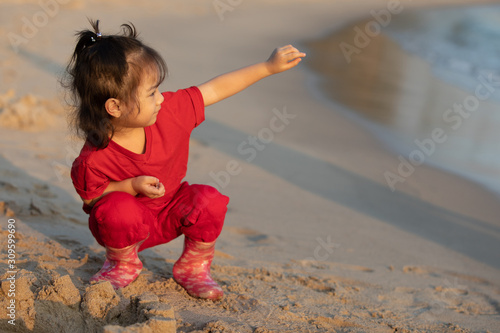A cute young girl, about 3 years old, in a bright red shirt, smiling, happy, playing on the sand. In the evening, by the beach, can see the beautiful sea.
