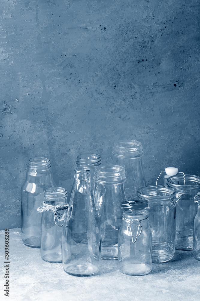 Variety of different shape empty opened glass bottles with and without lids standing on grey table with wall as background. Color of the year 2020 classic blue toned Variety of different shape empty opened glass bottles with and without lids standing on grey table with wall as background. Color of the year 2020 classic blue toned