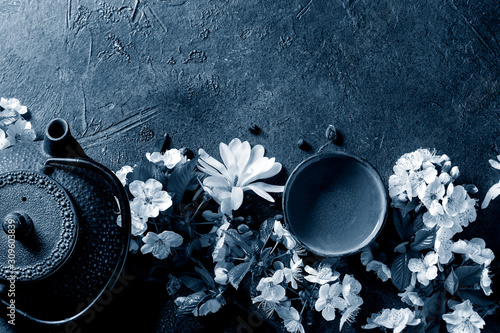 Traditional ceramic cup of hot tea with iron teapot, spring flowers magnolia and cherry blooming branches over dark texture background. Top view, copy space. Color of the year 2020 classic blue toned