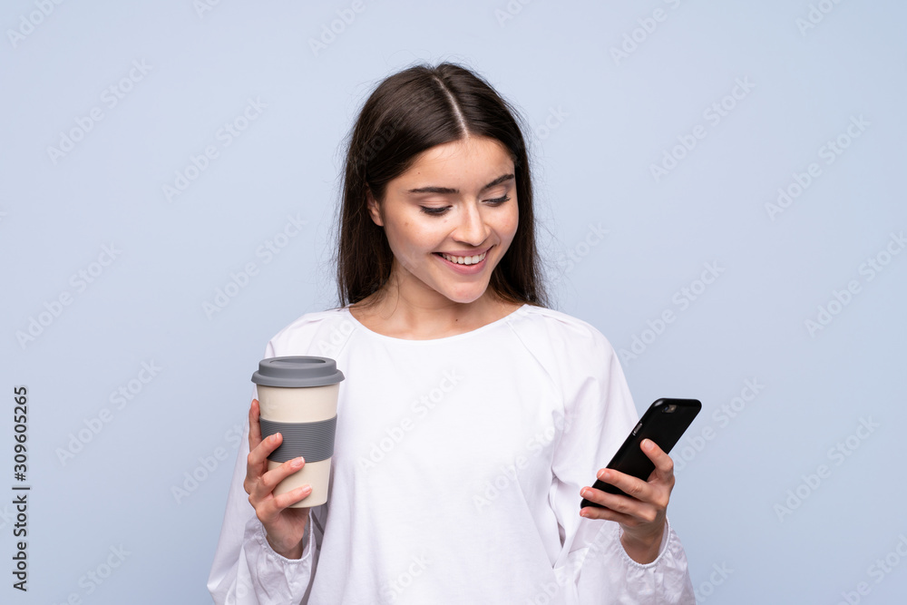 Young woman over isolated blue background holding coffee to take away and a mobile Young woman over isolated blue background holding coffee to take away and a mobile