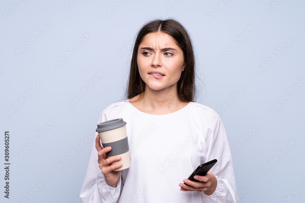 Young woman over isolated blue background holding coffee to take away and a mobile while thinking something Young woman over isolated blue background holding coffee to take away and a mobile while thinking something