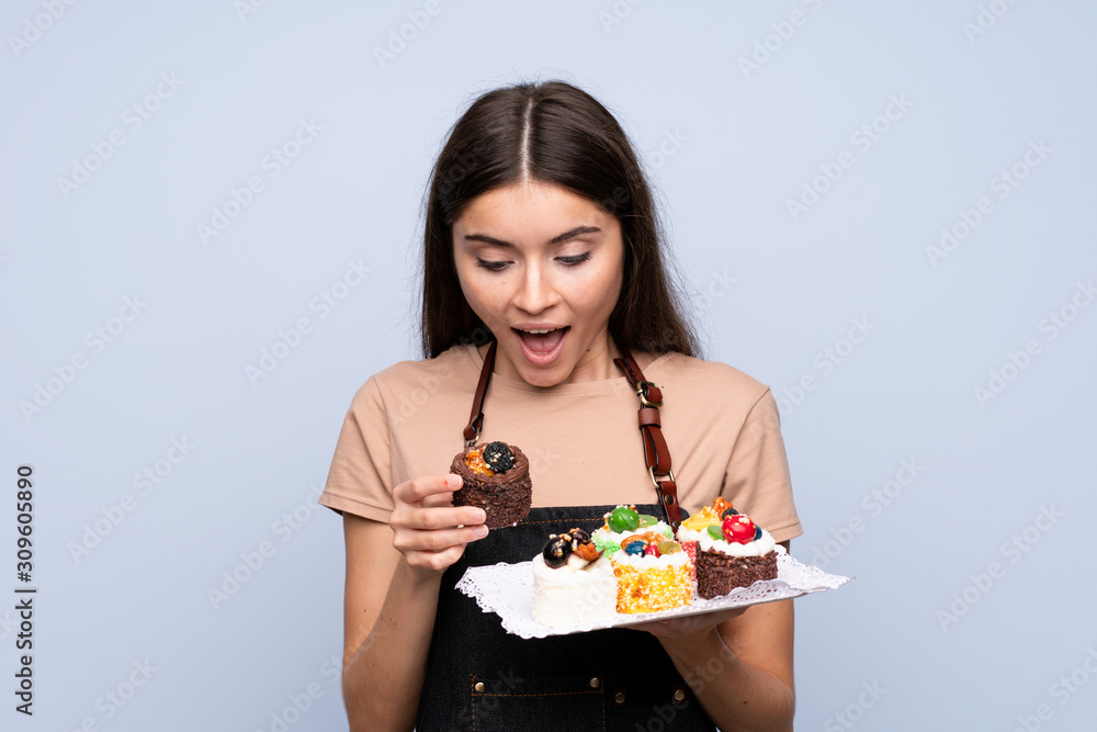 Young woman over isolated blue background holding mini cakes and surprised