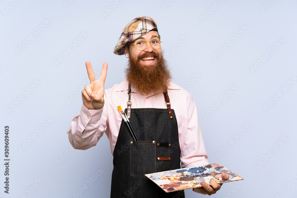 Redhead artist man with long beard holding a palette over isolated blue background smiling and showing victory sign