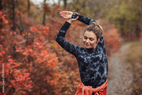 Young attractive sport woman in sportswear doing stretching exercise for hand in autumn forest with reddened leaves of trees. Warm up before training. Healthy lifestyle concept