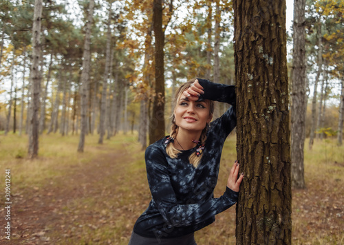 Young tired fit woman in sportswear leaned on a tree after an intense jog in the autumn forest. Healthy lifestyle concept. Outdoor workout